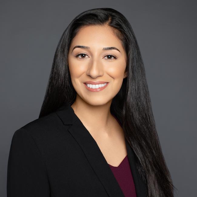 A headshot of Adrianna Carrasco, who is pictured in a maroon blouse and back blazer against a grey background. She is smiling, and has long dark hair.