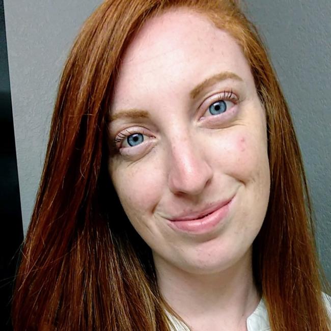 Headshot of Jessie Buth, a person with long red hair and blue eyes, smiling against a gray background, wearing a white top.