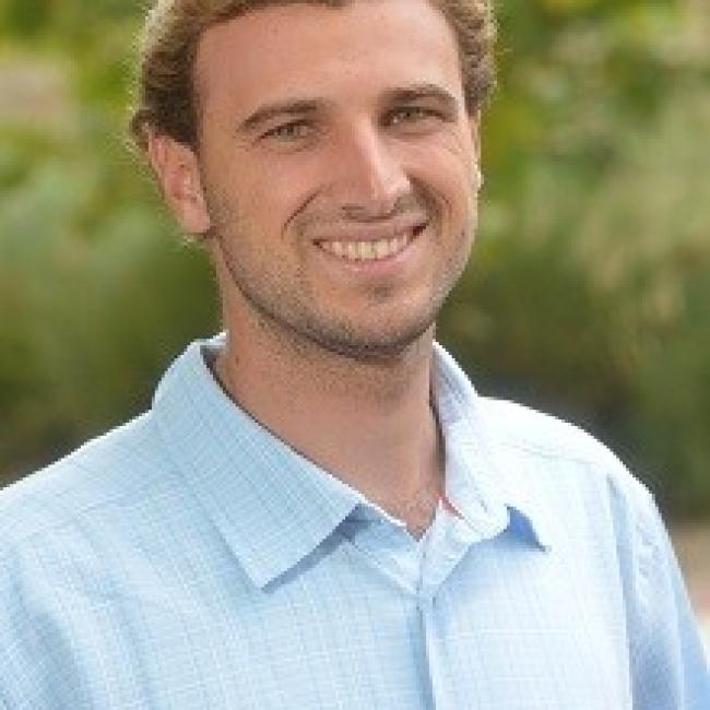 Headshot of Preston Crowell, a person with short blonde hair, smiling outdoors against a blurred green background, wearing a light blue button-up shirt.