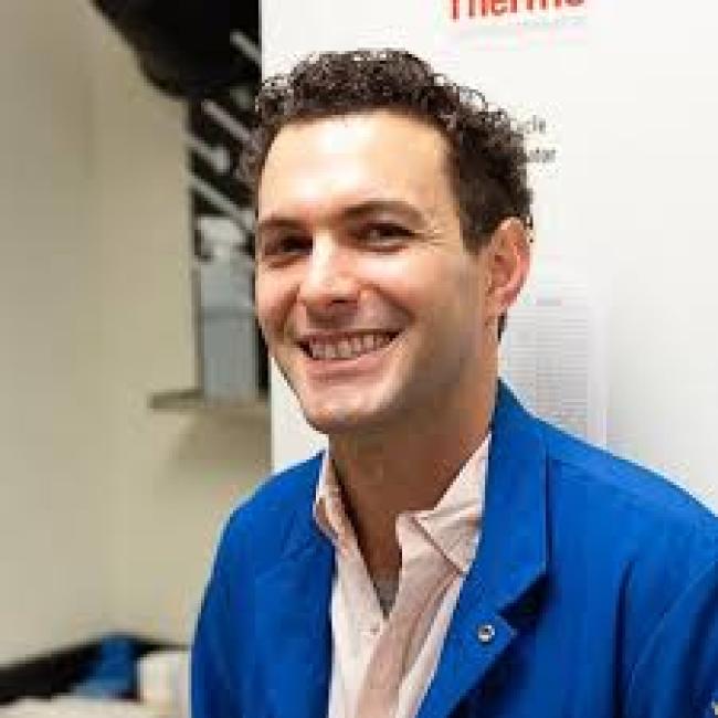 Headshot of Michael Hicks, a person with short dark curly hair, smiling in an indoor setting, wearing a blue lab coat over a light pink shirt.