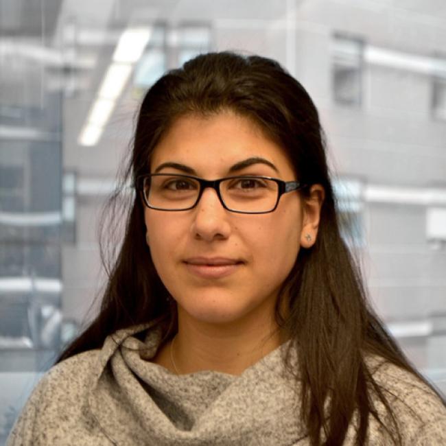 Headshot of BSCRC trainee Iman Fares, an individual with long dark hair and glasses wearing a textured gray top, indoors.