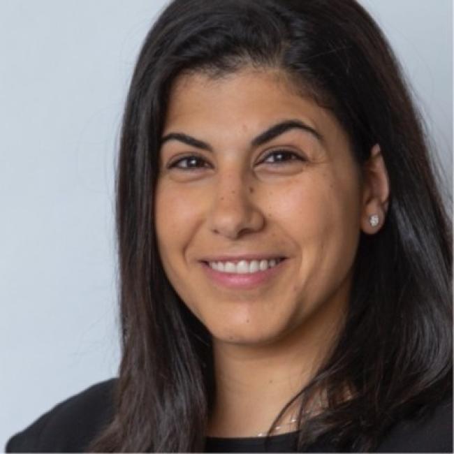 Headshot of Iman Fares, a person with long dark hair, smiling against a light gray background, wearing a dark top.