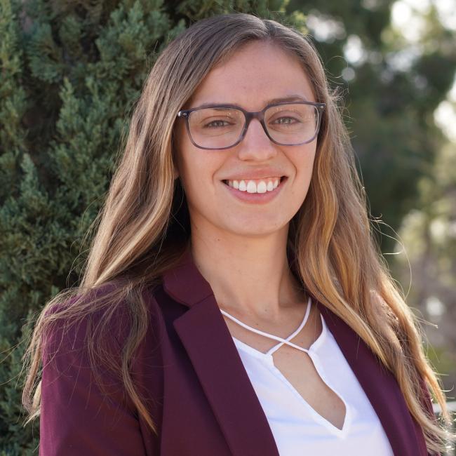 Headshot of Jenna Giafaglione, a woman with long light brown hair and glasses wearing a burgundy blazer, smiling outdoors.