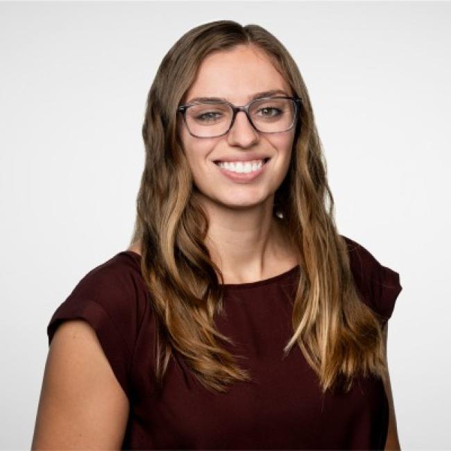 Headshot of Jenna Giafaglione, a person with long brown hair and glasses, smiling against a light gray background, wearing a dark maroon top.