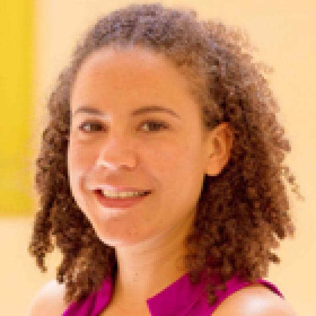  Headshot of Joanna Gell, short curly hair, smiling against a warm yellow background, wearing a bright pink top.