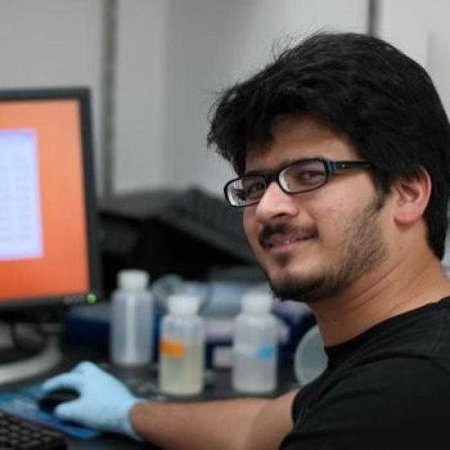 Headshot of Alok Joglekar, a person with short dark hair and glasses, smiling while working at a computer in a laboratory setting, wearing a black shirt and blue gloves.