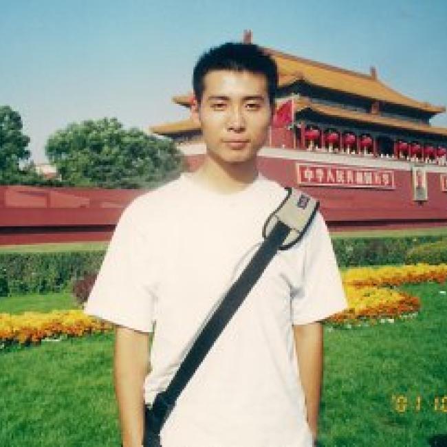 Headshot of Juehua Yu, a person with short dark hair, standing outdoors in front of a large red traditional building, wearing a white t-shirt and a shoulder bag.