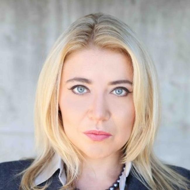 Headshot of Diana Katsman, a person with long blonde hair and blue eyes, against a light textured background, wearing a dark blazer and beaded necklace.