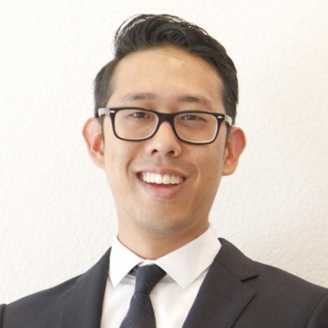 Headshot of William Kim, a person with short dark hair and glasses, smiling against a light background, wearing a dark suit jacket and tie.