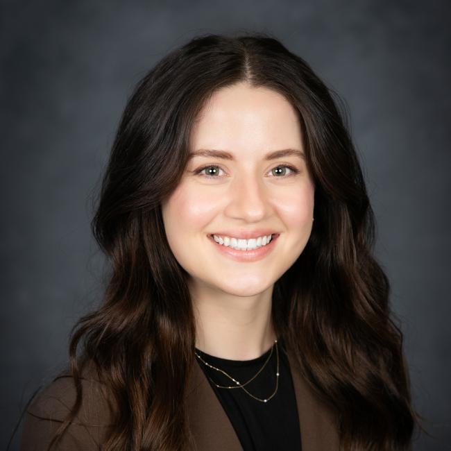 A headshot of Lauren Burnett, who is pictured against a dark background wearing a black shirt and a brown blazer. She has long brown hair and is smiling. 