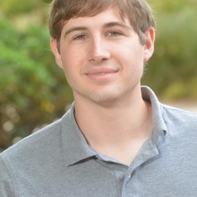 Headshot of Matthew Lowe, a young man with short brown hair wearing a gray polo shirt, smiling outdoors against a blurred green background.