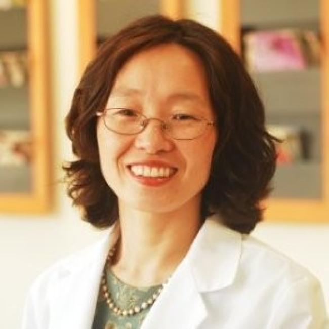 Headshot of Lydia Lee, a person with short dark hair and glasses, smiling in an indoor setting, wearing a white lab coat and pearl necklace.