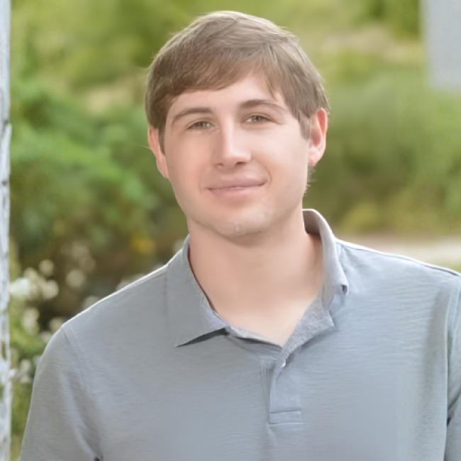 Headshot of Matthew Lowe, a young man with short brown hair wearing a gray polo shirt, smiling outdoors against a blurred green background.