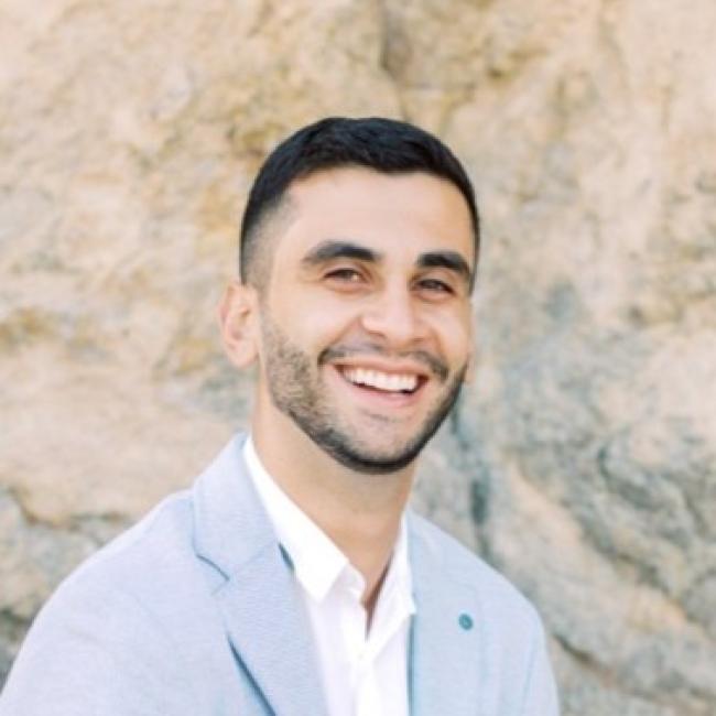 Headshot of Paul Ayoub, a person with short dark hair and a beard, smiling against a rocky outdoor background, wearing a light blue blazer and white shirt.