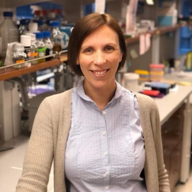 Headshot of Caroline Pearson, a person with short dark hair, smiling in a laboratory setting with lab equipment visible in the background, wearing a light blue shirt and tan cardigan.