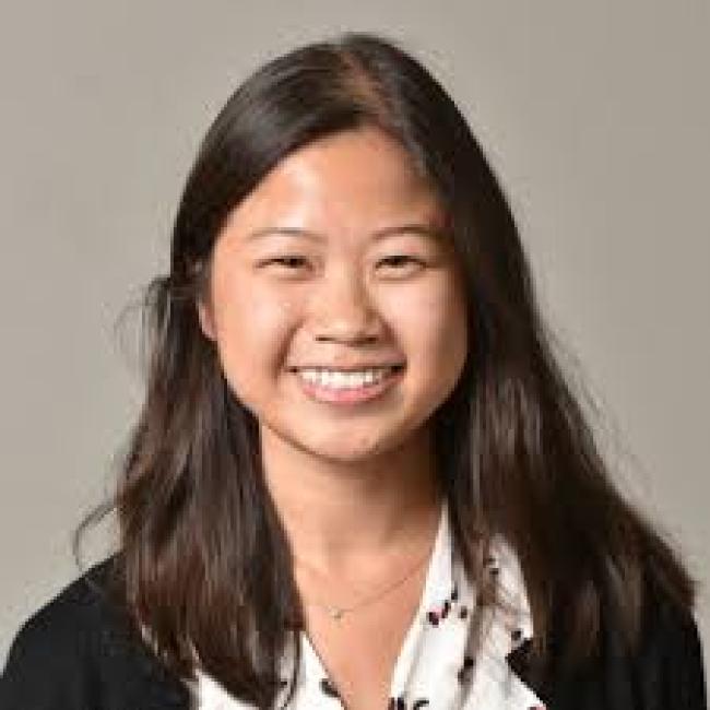 Headshot of Peggie Chien, a person with long dark hair, smiling against a gray background, wearing a black jacket and patterned top.