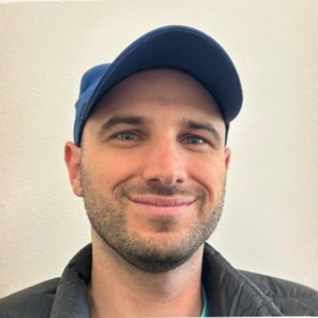 A close-up headshot of Preston Crowell smiling, wearing a blue baseball cap and a dark jacket against a plain white background.