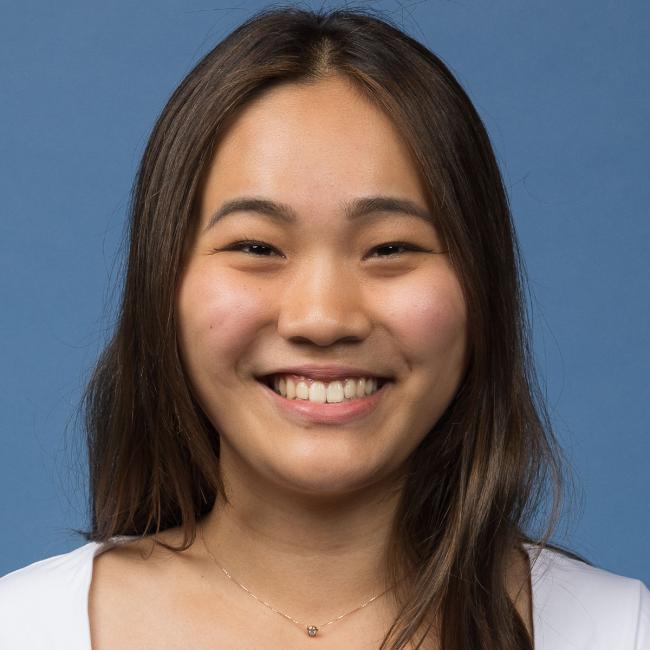 A headshot of Qui Ya Wu, who is smiling and is wearing a white top against a blue background. She also has long brown hair.