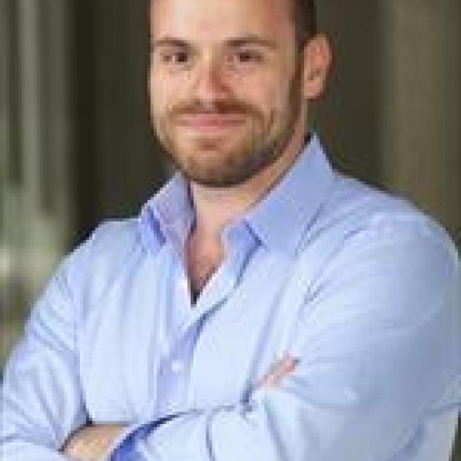 Headshot of Robert Singer, a person with short dark hair and a beard, smiling with arms crossed in an indoor setting, wearing a light blue shirt.