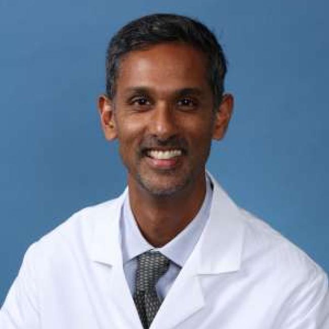 Headshot of Ranmal Samarasinghe, a person with short dark hair, smiling against a blue background, wearing a white lab coat with a UCLA Health logo and a gray patterned tie.