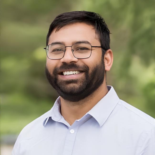 A headshot of Sandeep Gupta smiling, wearing glasses and a light-colored button-up shirt, with a blurred green outdoor background.