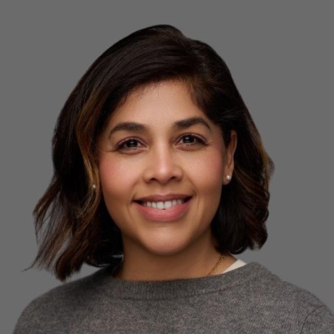 Headshot of Salemiz Sandoval, a person with shoulder-length dark hair, smiling against a gray background, wearing a gray top.