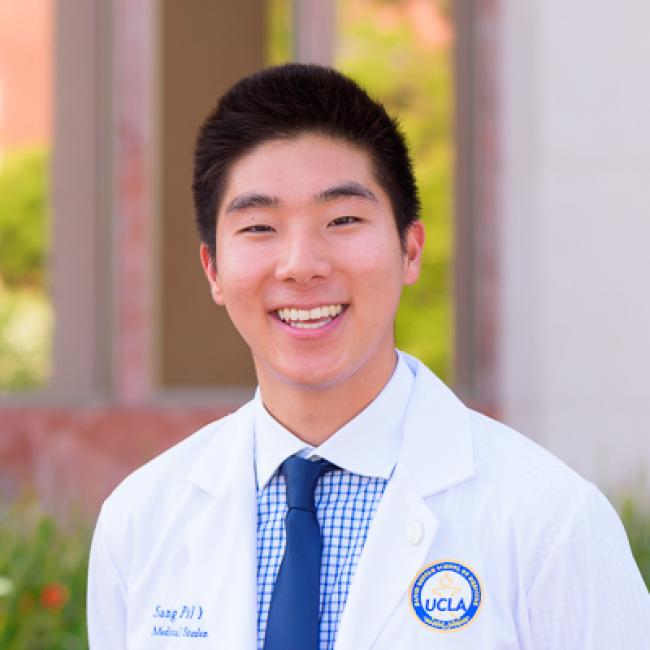 Headshot of Sang-Pil (Alex) Yoo, a person with short dark hair, smiling outdoors against a blurred background, wearing a white UCLA lab coat and blue checked shirt with a navy tie.