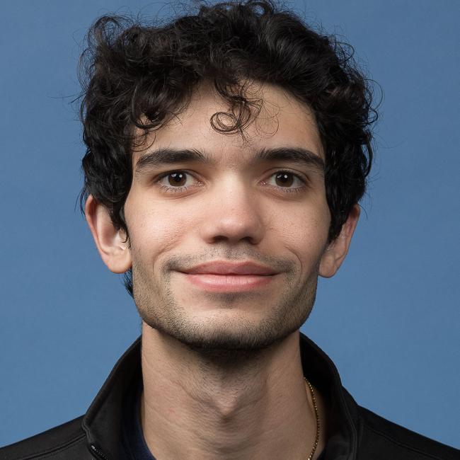 A headshot of Satchel Stevens, who is dressed in a black button-up shirt and is smiling against a blue background. 