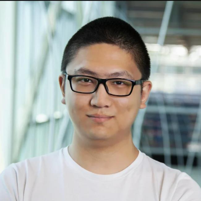 Headshot of Xuegang Yuan, a person with short dark hair and black-framed glasses, smiling in a modern indoor setting, wearing a white t-shirt.