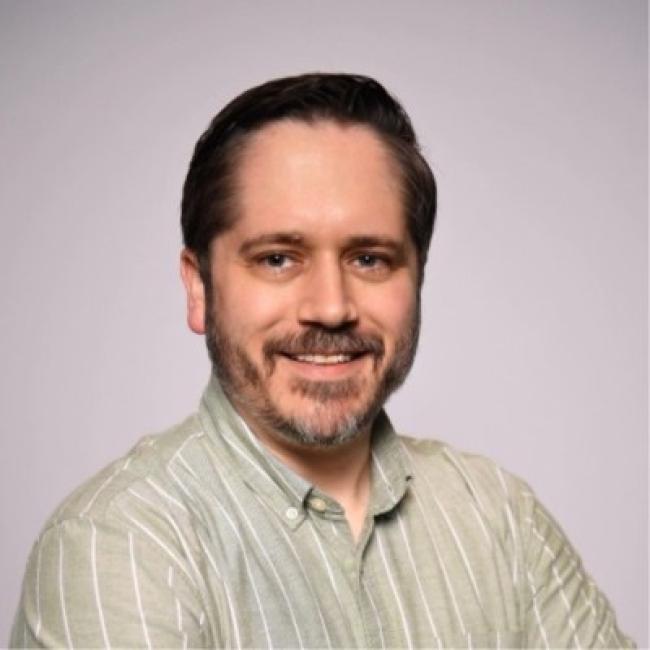 Headshot of Sean Sherman, a person with short dark hair and a beard, smiling against a light gray background, wearing a striped green shirt.