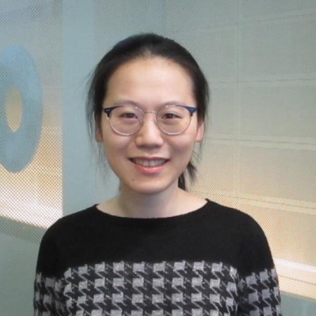 A headshot of Shang-Jung Cheng, who stands smiling in glasses and a black-and-white sweater. She is standing indoors against a white background. 