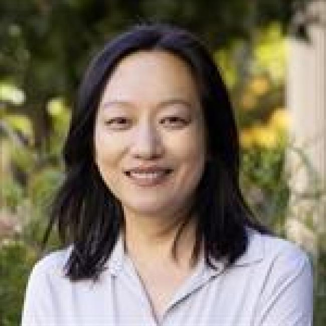 Headshot of Yin Shen, a person with shoulder-length dark hair, smiling outdoors against a blurred green background, wearing a light-colored top.