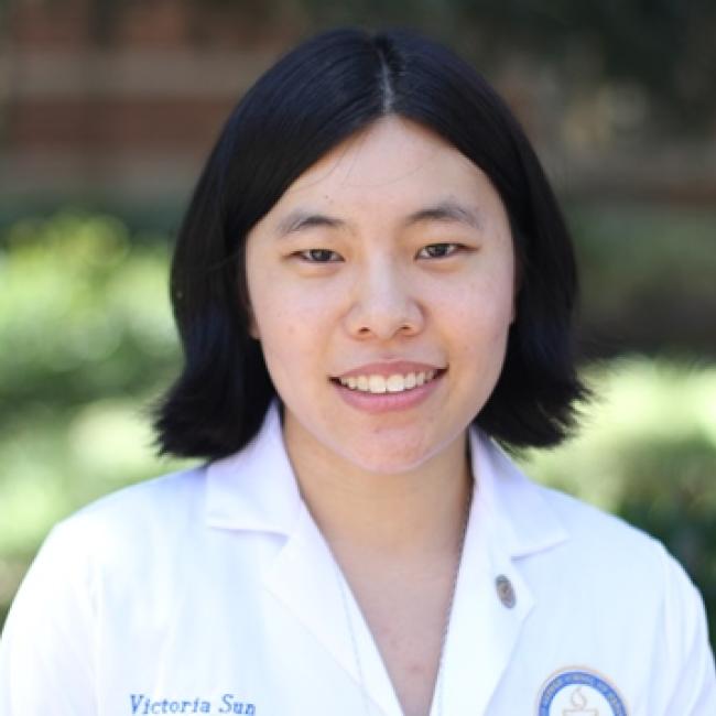 Headshot of Victoria Sun, a person with short dark hair, smiling outdoors against a blurred green background, wearing a white lab coat.