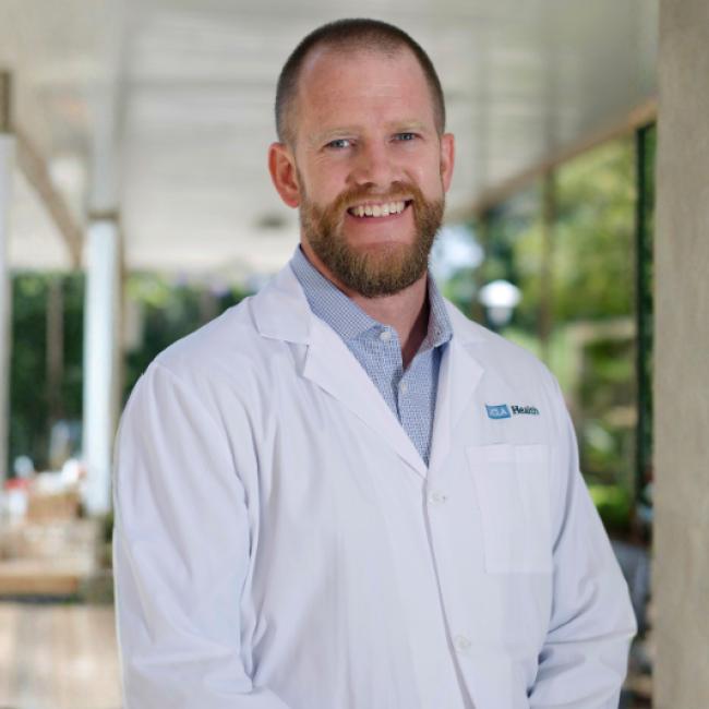 UCLA researcher Vondriska, Thomas smiles for a photograph. He wears a white UCLA labcoat and stands indoors with a green landscape appearing behind him.