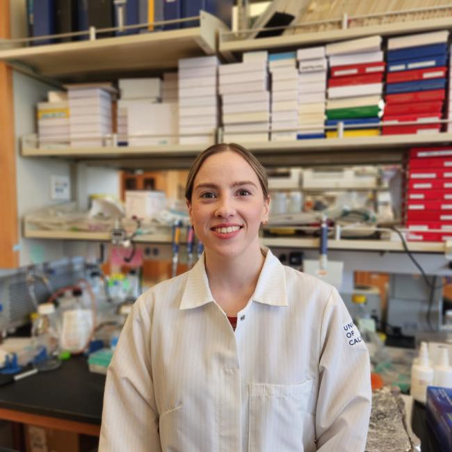 Headshot of Anastasia Vavilina-Halstead, a person with dark hair pulled back, smiling in a laboratory setting with shelves of binders and lab equipment in the background, wearing a University of California lab coat.