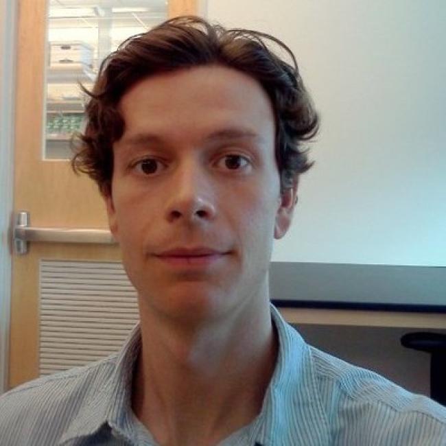 Headshot of Wade Richardson, a person with short wavy dark hair, in an indoor setting with a wooden door visible in the background, wearing a striped shirt.