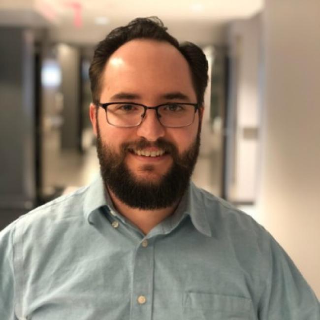 Headshot of Dan Wilkison, a person with short dark hair, a beard, and glasses, smiling in an indoor hallway setting, wearing a light blue button-up shirt.