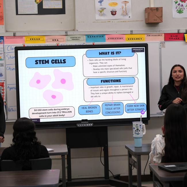 two female researchers stand in front of a high school classroom with a TV monitor in between them projecting a slide on stem cells