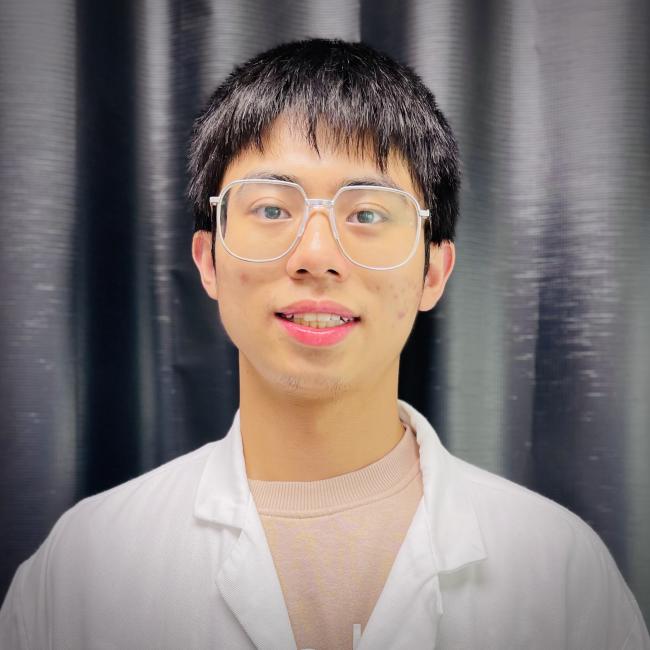 A headshot of Yichen Zhu, who stands smiling in a white lab coat and glasses against a background of black curtains. 
