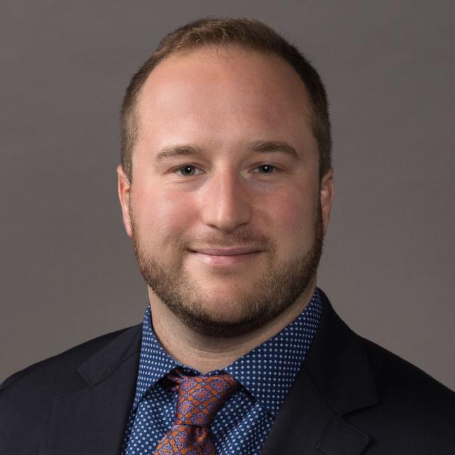 A headshot of Zachary Olmsted, who is pictured wearing a black blazer over a navy blue button-up shirt and a mahogany tie. 